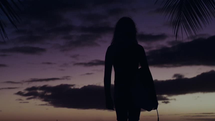 Silhouette of a female surfer holding her board, looking out at the colorful twilight sky