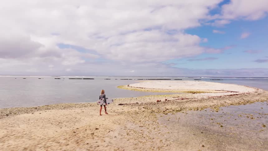 Aerial shot follows a young surfer carrying a surfboard while walking along a stunning sandbar in the middle of the ocean
