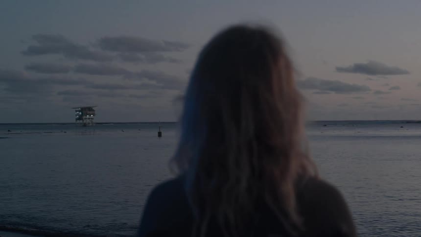 A contemplative shot from behind an out-of-focus woman looking at an illuminated platform on the calm sea during twilight