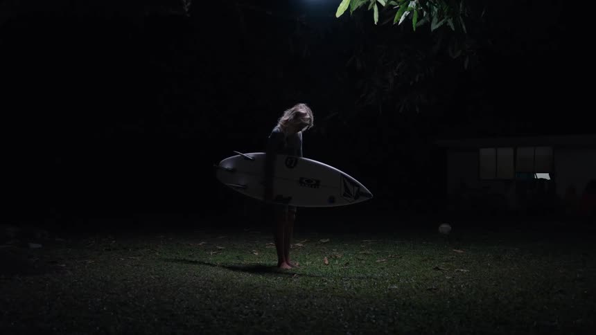 A lone surfer stands under a spotlight in a dark yard at night, holding her surfboard