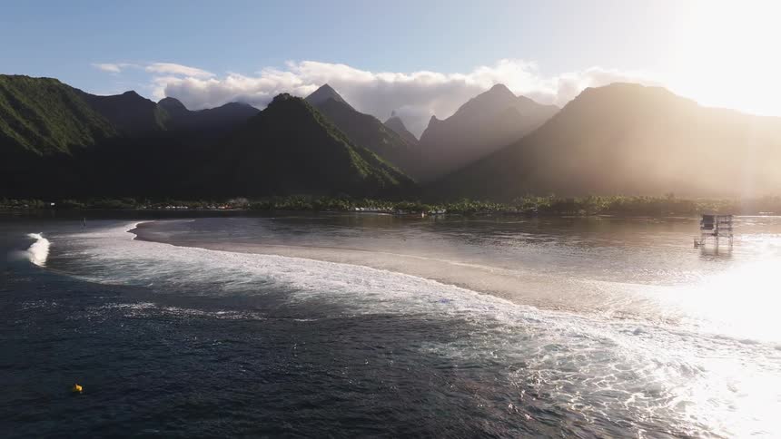 Golden sunlight illuminates the iconic Teahupo'o surf break in Tahiti as waves roll in with lush green mountains in the background
