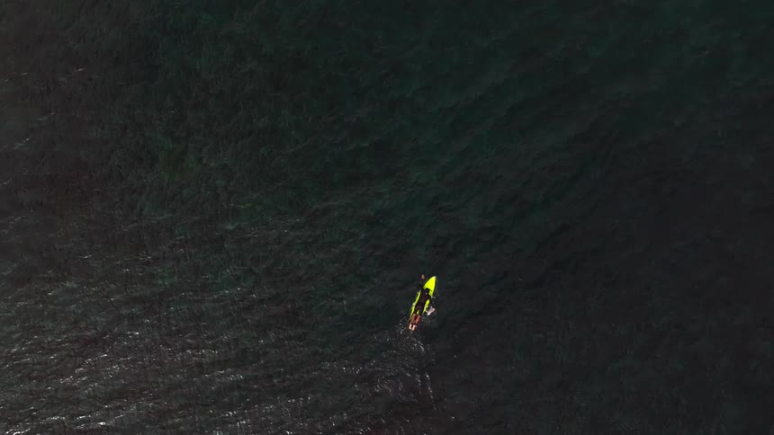An aerial top-down view shows a lone surfer paddling on a yellow board across the vast, dark ocean water