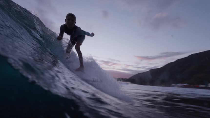 A talented young girl skillfully surfs a dark ocean wave in stunning slow motion as the sun sets, with the camera capturing the action from just above and below the water