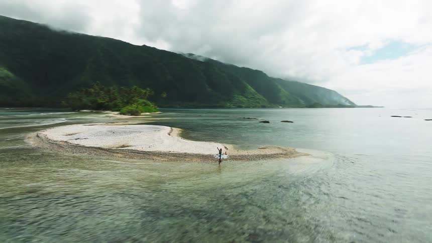 An epic drone shot flies towards a lone surfer on a remote sandbar, revealing a stunning tropical island with lush green mountains and a clear turquoise lagoon