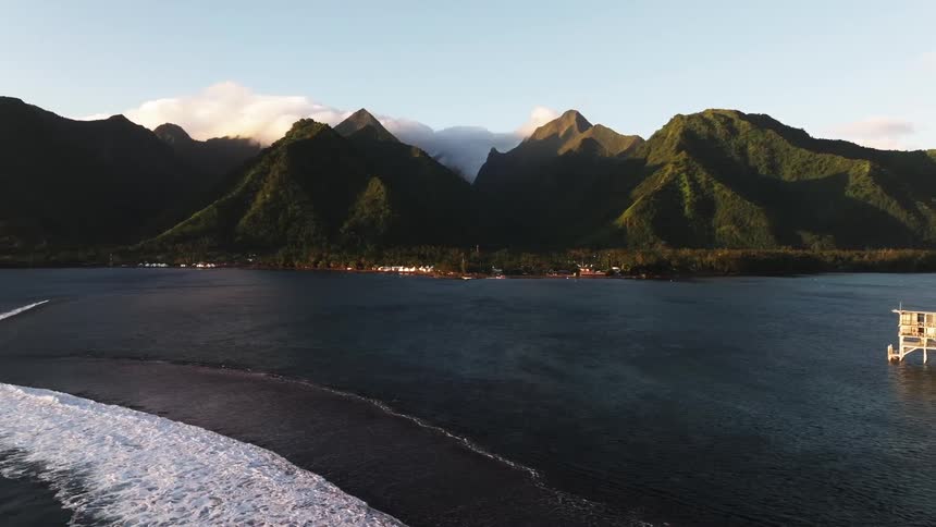 An aerial view reveals a stunning tropical coastline with breaking waves, a seaside village, and a stilted house set against a backdrop of majestic green mountains