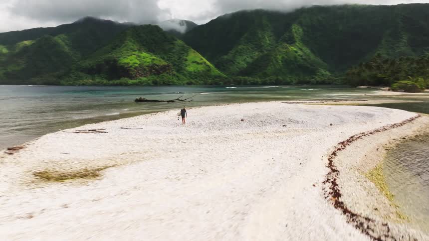 An aerial view follows a surfer as she walks with her board across a secluded white sandbar, backed by lush green tropical mountains