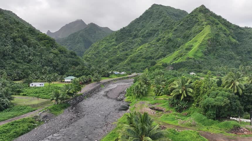 A stunning aerial view pulls back over a river running through a lush green tropical valley surrounded by towering mountains on a cloudy day