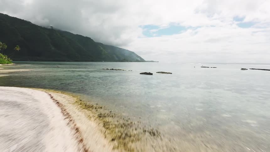 Aerial drone flies low over a shallow tropical lagoon past a woman standing on a remote sandbar with lush mountains