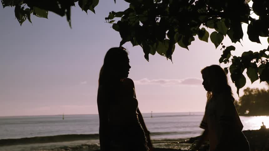 Silhouettes of a mother and daughter playfully interacting under a tree on a tropical beach at sunset
