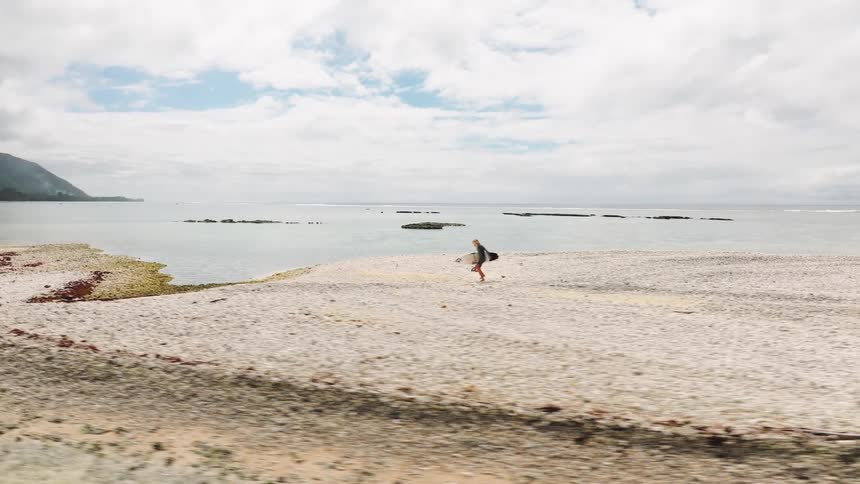 An aerial follow shot shows a woman carrying her surfboard as she walks along a stunning rocky beach on a tropical island