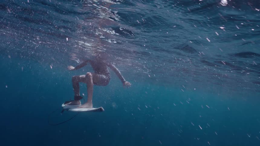 A serene slow-motion underwater shot captures a surfer waiting on their board in the deep blue ocean