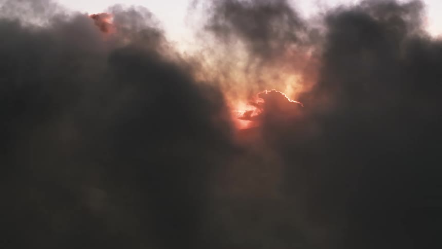 Thick black smoke billows across the sky, almost completely obscuring the fiery sun