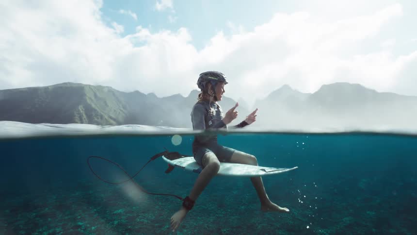 A surfer in a helmet waits on his board in crystal clear turquoise water, captured in a slow-motion over-under shot with lush tropical mountains behind him