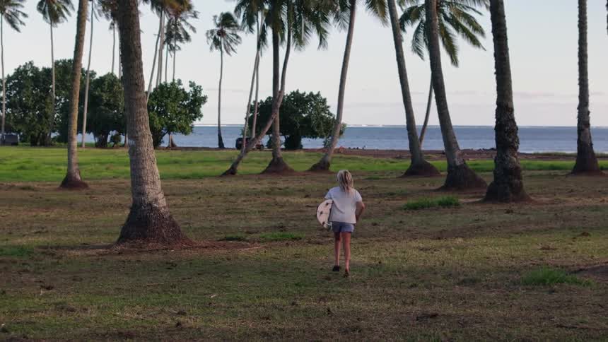 A young surfer eagerly runs with their board through a grassy palm tree grove on their way to the ocean in slow motion