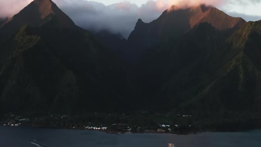 A small boat sails along a tropical island coastline at dawn, with the camera tilting up to reveal majestic mountains catching the first light of day