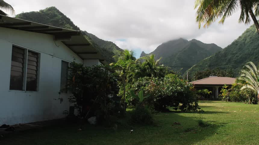 A peaceful tropical home with chickens on the lawn sits at the base of lush, green mountains