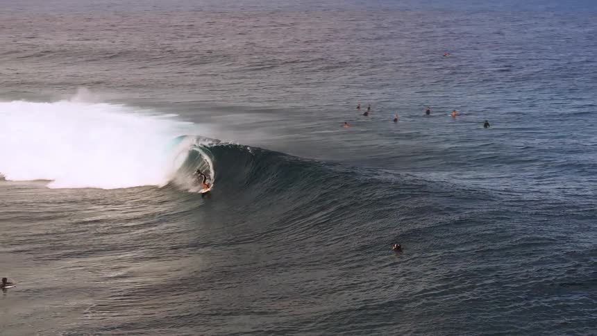 A lone surfer rides the barrel of a massive, powerful ocean wave in dramatic slow motion as other surfers watch