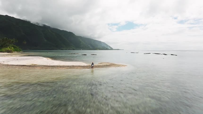 An aerial view flies over a lone woman standing on a remote sandbar in the crystal clear waters of a tropical island