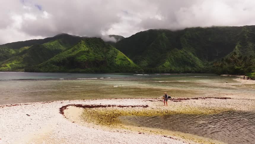 An aerial view shows a lone surfer walking with her surfboard along a white beach on a remote tropical island with lush green mountains in the background