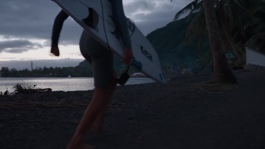 A surfer in a wetsuit walks barefoot with her surfboard along a dark, tropical beach at dusk