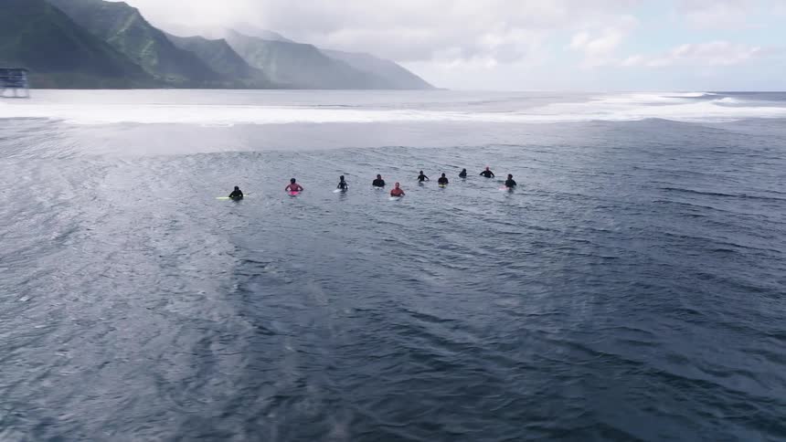 Aerial view of a group of surfers patiently waiting for waves in the ocean with a stunning mountainous coastline in the background