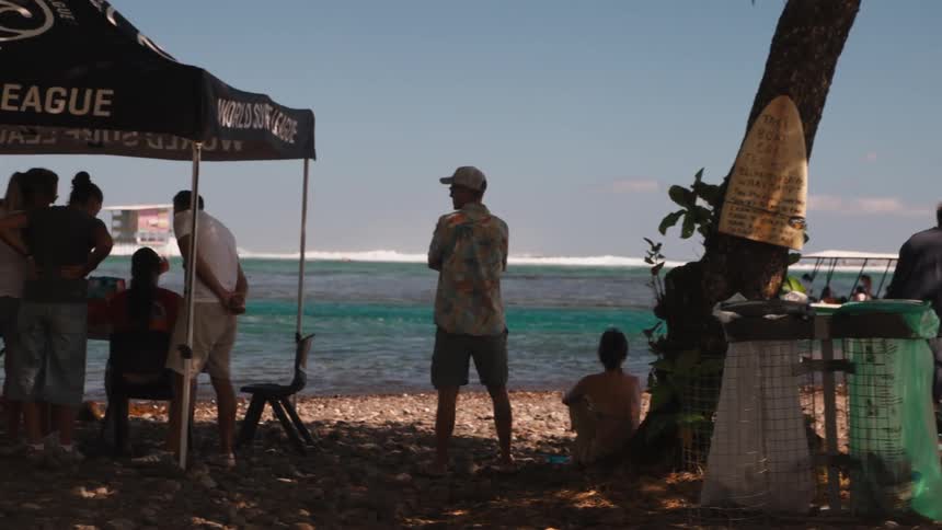 Spectators gather on a tropical beach under a World Surf League tent, watching a competition on the turquoise ocean