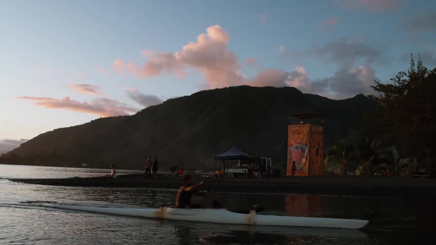 A person in an outrigger canoe paddles in slow motion at dusk near a beach with mountains in the background