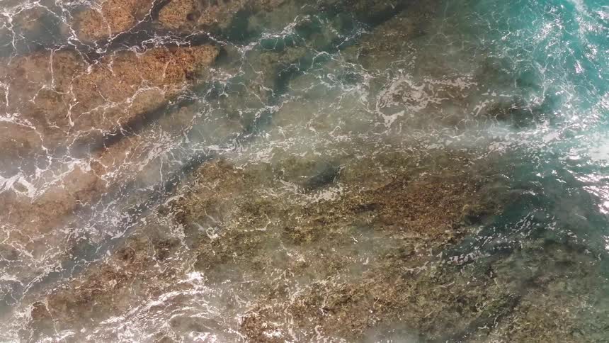 An aerial top-down view captures a powerful turquoise ocean wave breaking over a shallow reef in dramatic slow motion
