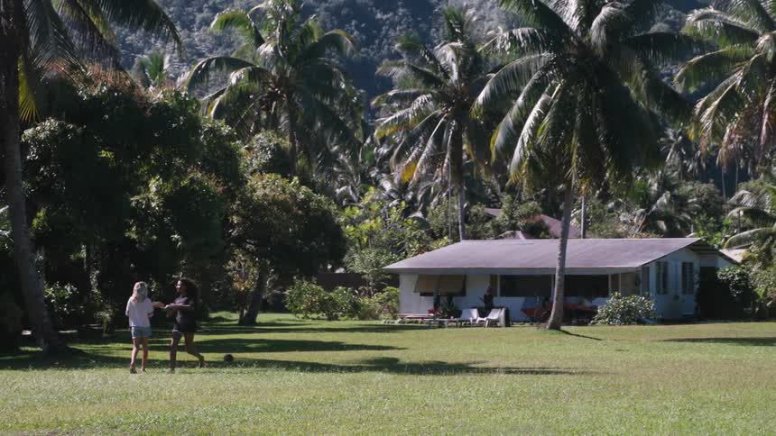 Two female friends run to each other and embrace in a slow-motion hug on a grassy lawn surrounded by palm trees