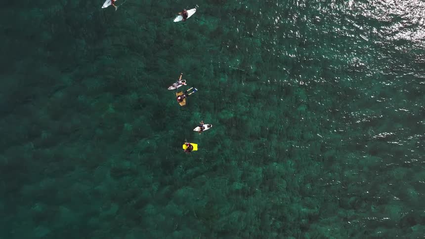 A serene top-down aerial view of a group of surfers relaxing on their boards in the calm, clear turquoise ocean