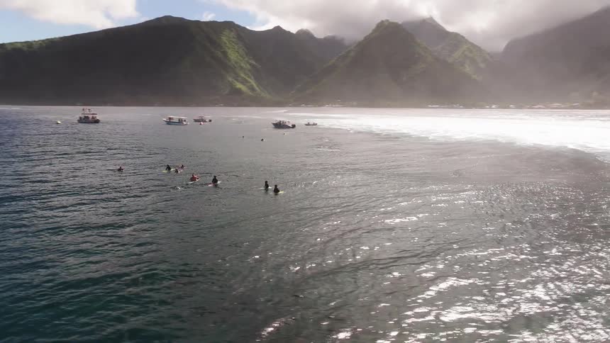 An expansive aerial shot reveals a group of surfers waiting patiently in the deep blue ocean near a breathtakingly lush and mountainous tropical coastline