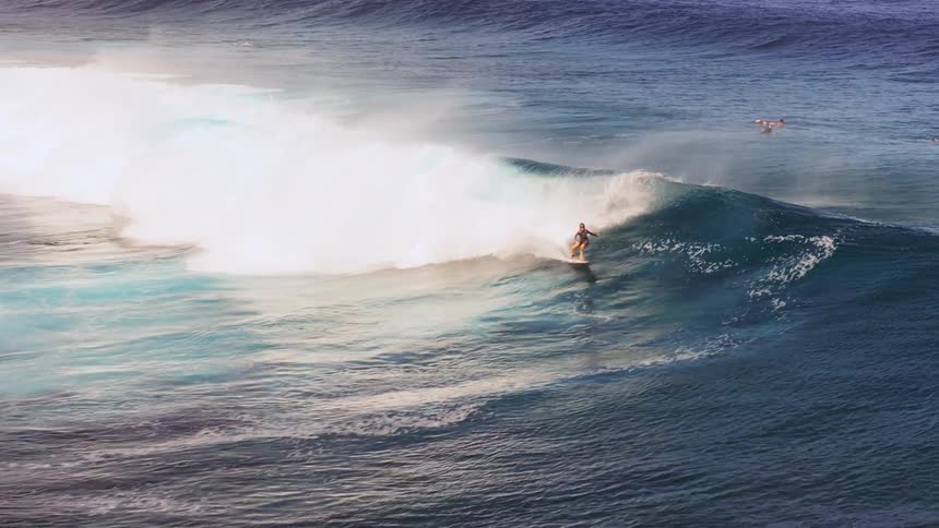 Slow motion footage shows a skilled surfer riding a massive, barreling ocean wave as other surfers look on