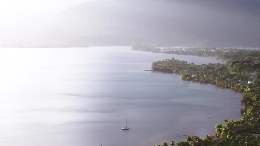 A bright mist slowly envelops a tranquil tropical bay with a lone sailboat and lush coastal village