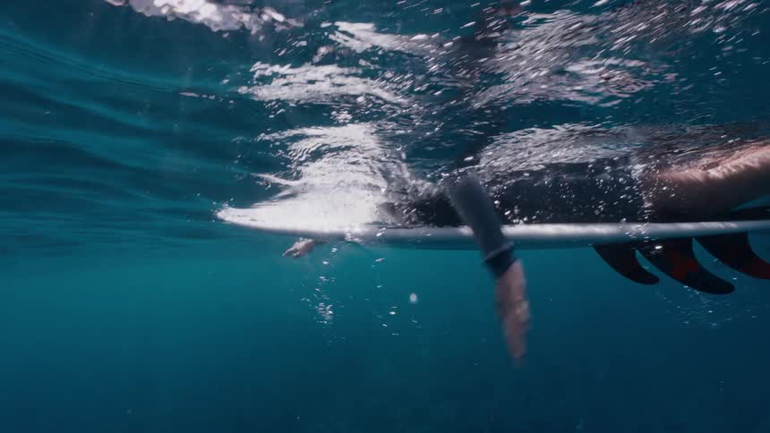 Underwater slow-motion view of a surfer paddling on their surfboard in clear blue ocean water