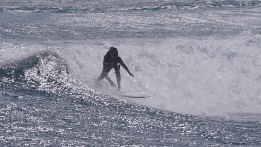 A skilled female surfer in a wetsuit gracefully rides a sunlit ocean wave, performing a powerful cutback in super slow motion