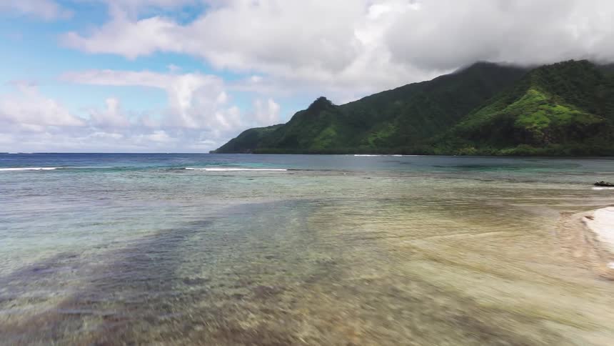 Aerial drone shot flying over a crystal clear tropical lagoon with a visible coral reef and sandbar towards a lush green mountainous island