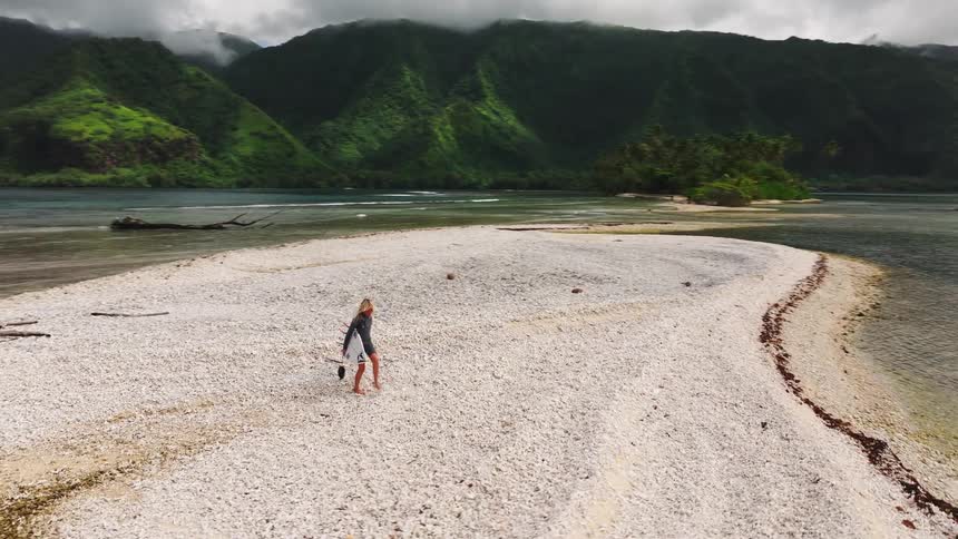 A female surfer walks along a secluded coral beach spit, surfboard in hand, surrounded by the clear ocean and backed by verdant tropical mountains