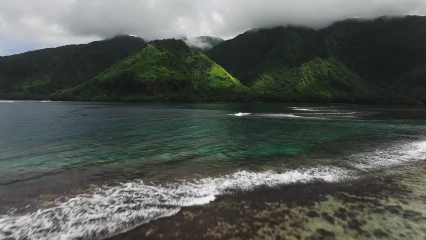 An aerial shot flies over a woman carrying her surfboard along a tropical sandbar towards the ocean, with lush green mountains in the background