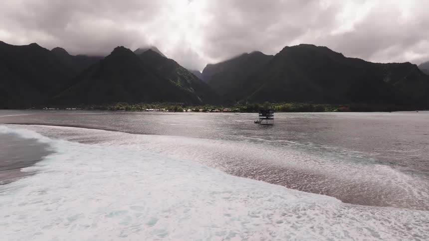 A dramatic aerial slow-motion shot glides over a breaking ocean wave towards a judging tower, with the lush, cloudy mountains of Tahiti in the background