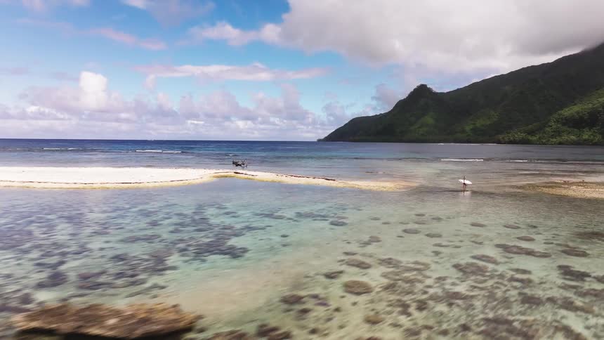 Stunning aerial footage glides over a shallow coral reef and turquoise lagoon towards a lone surfer on a tropical island