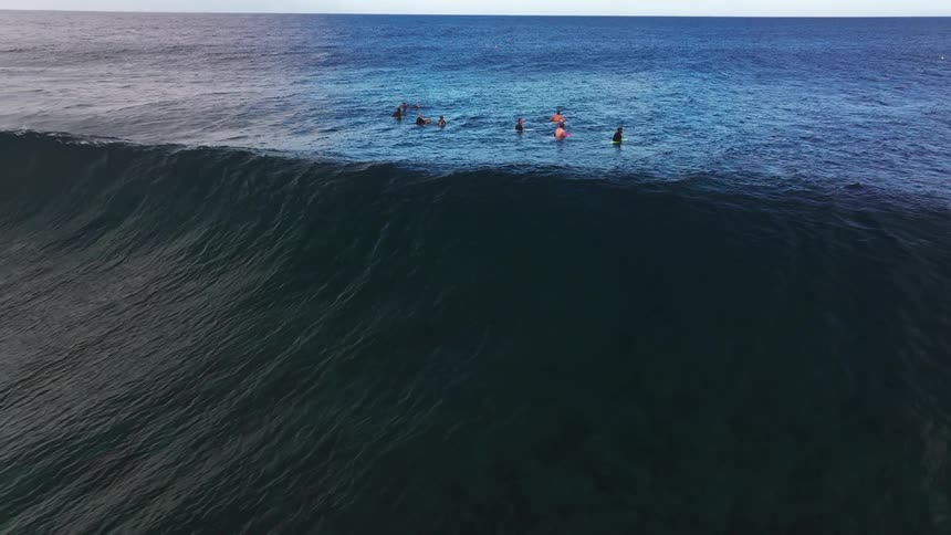 Dramatic slow-motion aerial shot of surfers waiting in the deep blue ocean as a massive, dark wave builds and approaches