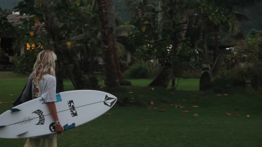 A young woman with a surfboard walks from her house through a tropical yard to a pickup truck, ready for a day of surfing