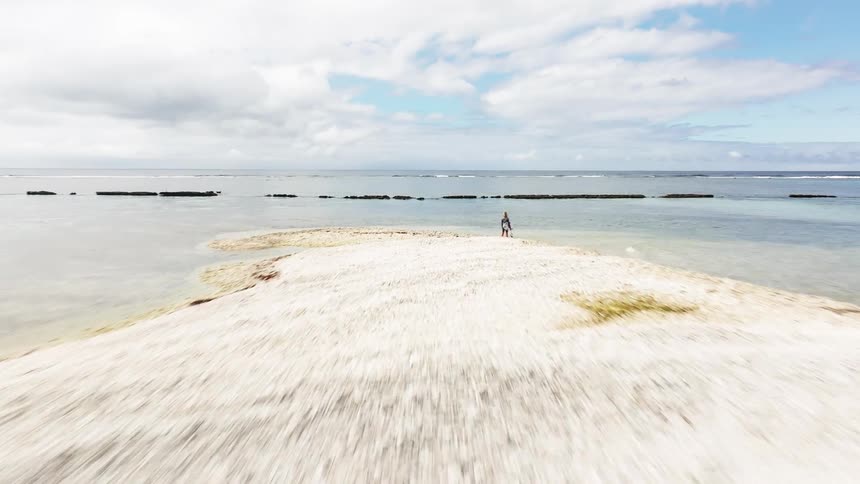 A fast-moving aerial shot flies low over a clear turquoise ocean, passing a small sandbar with a lone person standing on it towards the open sea