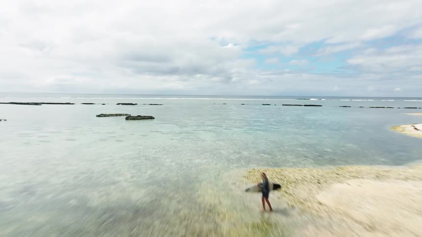Aerial drone shot flies over a tropical sandbar with a surfer towards the clear turquoise ocean and coral reef