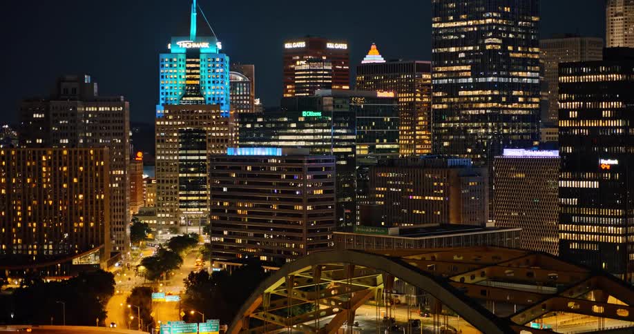 Aerial view of the illuminated downtown Pittsburgh skyline at night with the iconic yellow bridge and city streets in the foreground