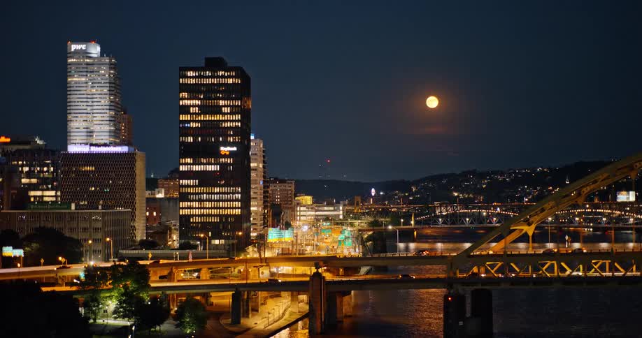 A brilliant full moon rises over the illuminated downtown Pittsburgh city skyline at night as traffic crosses the bridges over the river
