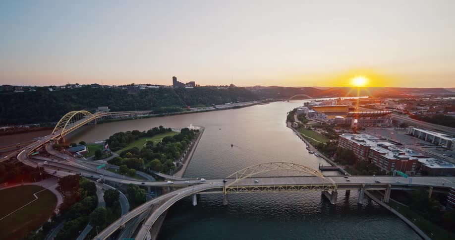 A beautiful golden hour aerial view of Pittsburgh's Point State Park where the rivers meet, with cars crossing the bridges at sunset