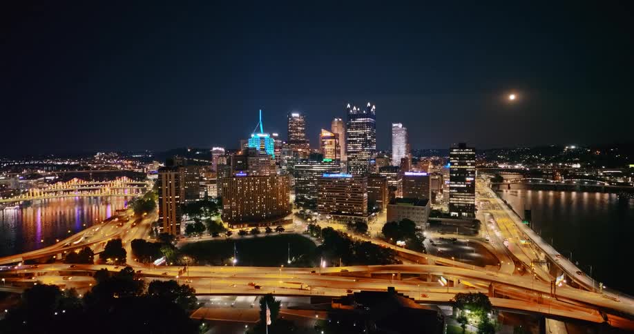 An aerial view pulls back to reveal the glowing Pittsburgh city skyline at night, with traffic streaming along the highways and bridges