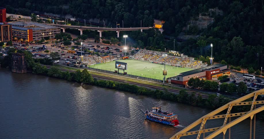 An aerial view of a soccer game at a riverside stadium at dusk as a riverboat and train pass by