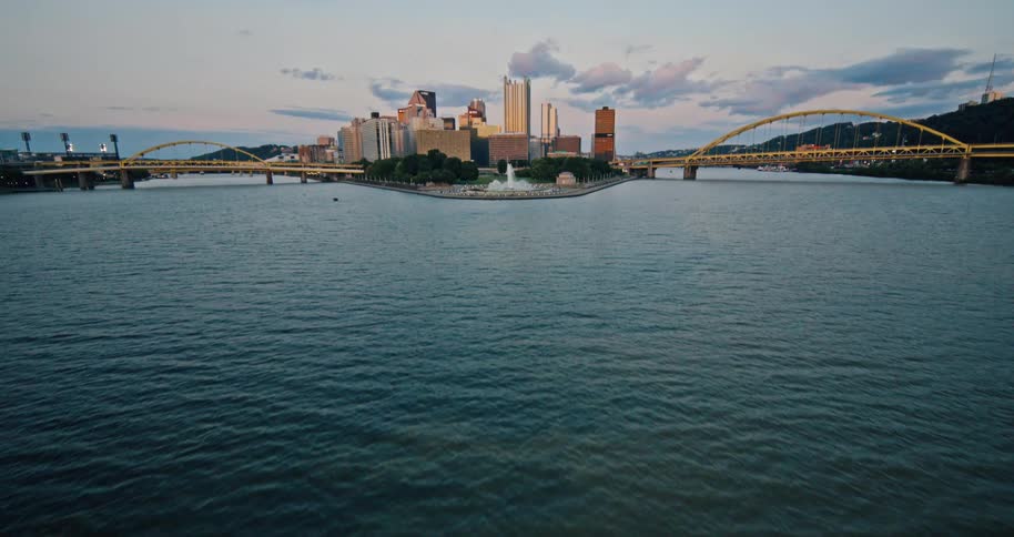 An aerial shot tilts up from the river to reveal the iconic Pittsburgh, Pennsylvania skyline and its famous yellow bridges at sunset
